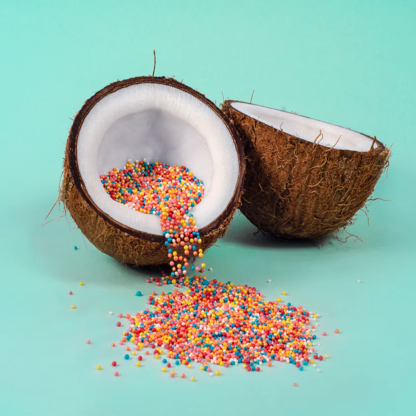 An open coconut with multi-coloured sweets spilling out of one half. Set on a teal background.