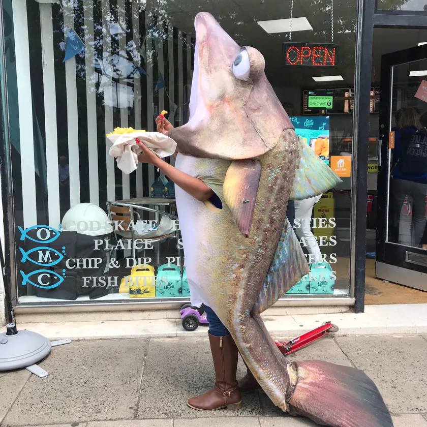 A person in a fish costume standing outside a shop eating fish and chips